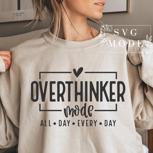 May include: A beige sweatshirt with the words "OVERTHINKER mode ALL &bull; DAY &bull; EVERY &bull; DAY" in black block letters. A black heart is above the word "OVERTHINKER". The sweatshirt is a neutral color and has long sleeves.