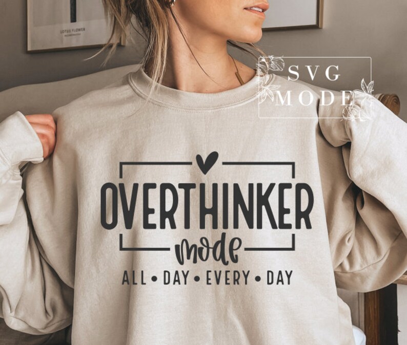 May include: A beige sweatshirt with the words "OVERTHINKER mode ALL &bull; DAY &bull; EVERY &bull; DAY" in black block letters. A black heart is above the word "OVERTHINKER". The sweatshirt is a neutral color and has long sleeves.