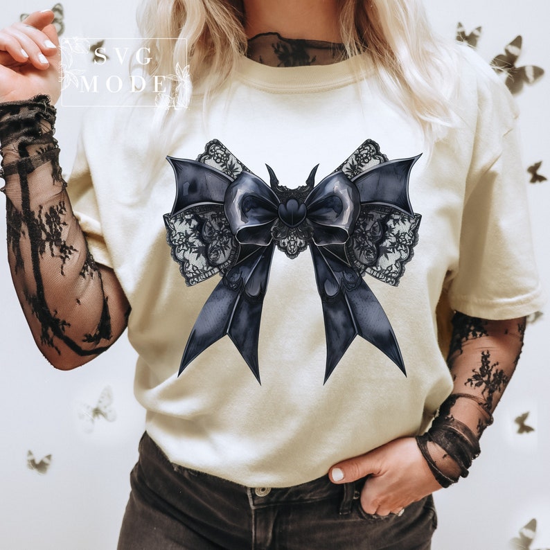 May include: A white t-shirt with a black and white lace bow with bat wings. The bow is a dark blue color and has a heart in the center.