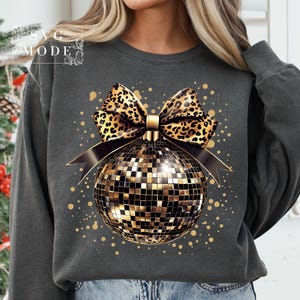 May include: A gray sweatshirt with a gold disco ball ornament and a leopard print bow. The ornament is surrounded by gold glitter.