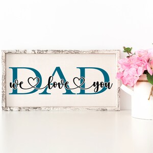 Puede incluir: Un letrero de madera blanco con la palabra "DAD" en letras azul turquesa. Las letras est&aacute;n conectadas por una escritura cursiva negra que dice "we love you" con un s&iacute;mbolo de coraz&oacute;n entre cada palabra.