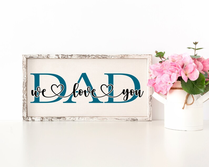 Puede incluir: Un letrero de madera blanco con la palabra "DAD" en letras azul turquesa. Las letras est&aacute;n conectadas por una escritura cursiva negra que dice "we love you" con un s&iacute;mbolo de coraz&oacute;n entre cada palabra.