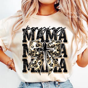 Puede incluir: Camiseta color crema con las palabras "Praying Mama" en escritura negra y letras de bloque. Un lazo con estampado de leopardo con una cruz est&aacute; centrado en la camiseta. La camiseta tiene mangas cortas y cuello redondo.
