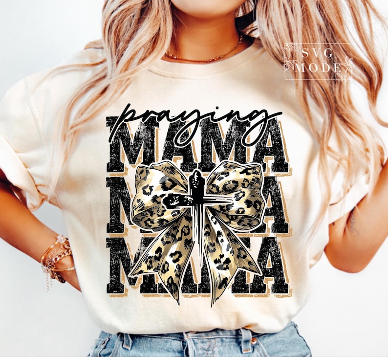 Puede incluir: Camiseta color crema con las palabras "Praying Mama" en escritura negra y letras de bloque. Un lazo con estampado de leopardo con una cruz est&aacute; centrado en la camiseta. La camiseta tiene mangas cortas y cuello redondo.