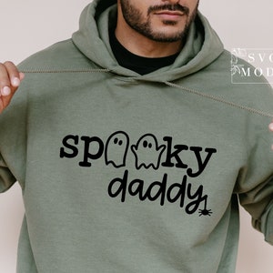 May include: A green hooded sweatshirt with the text "spooky daddy" in black lettering. Three ghost silhouettes are above the text. A small black spider is below the text.