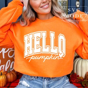 Może przedstawiać: Pomarańczowa bluza z białym napisem "HELLO pumpkin" z małym białym serduszkiem. Bluzę nosi osoba z blond włosami.