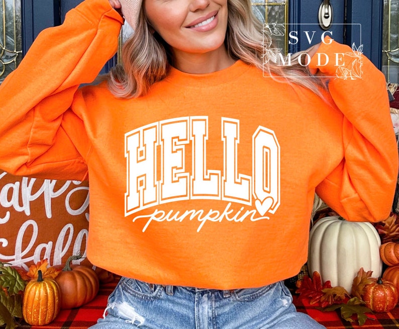Może przedstawiać: Pomarańczowa bluza z białym napisem "HELLO pumpkin" z małym białym serduszkiem. Bluzę nosi osoba z blond włosami.
