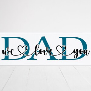 Puede incluir: Se&ntilde;al de madera blanca con la palabra "DAD" en letras azul turquesa. Las palabras "we love you" est&aacute;n escritas en cursiva negra con un coraz&oacute;n entre cada palabra.
