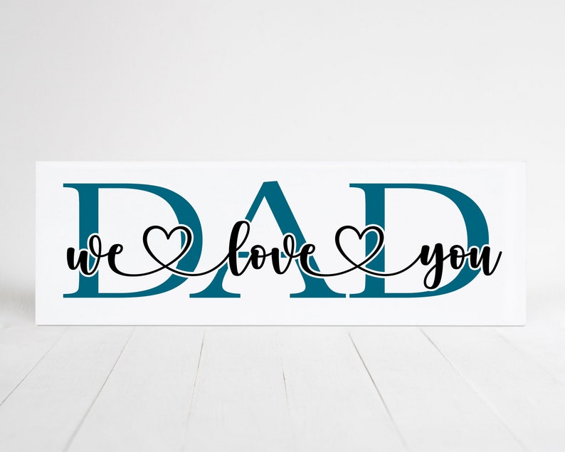 Puede incluir: Se&ntilde;al de madera blanca con la palabra "DAD" en letras azul turquesa. Las palabras "we love you" est&aacute;n escritas en cursiva negra con un coraz&oacute;n entre cada palabra.