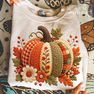 May include: A beige crewneck sweatshirt featuring a large, embroidered-style graphic of a pumpkin. The pumpkin is composed of various textured panels in orange, green, and cream, decorated with floral motifs, berries, and autumn leaves. Text reads SVG MODE.