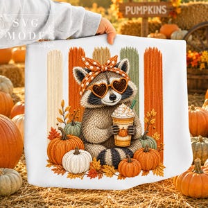 May include: A white sweatshirt featuring a graphic of a raccoon wearing a polka dot headband and heart-shaped sunglasses, holding a coffee cup. The raccoon is surrounded by pumpkins and autumn leaves against a background of vertical stripes.