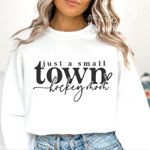 Op de afbeelding: Witte trui met zwarte tekst die "just a small town hockey mom" luidt met een klein hartje na "town".