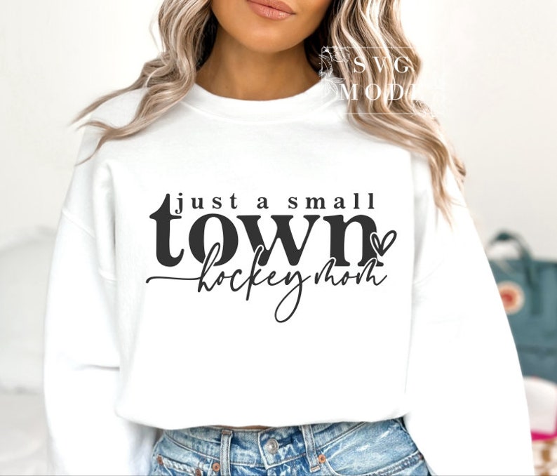 Op de afbeelding: Witte trui met zwarte tekst die "just a small town hockey mom" luidt met een klein hartje na "town".