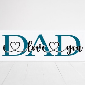 Puede incluir: Un letrero de madera blanco con la palabra "DAD" en letras azul turquesa. Las letras est&aacute;n conectadas por una l&iacute;nea negra con dos corazones. El texto "I love you" est&aacute; escrito en cursiva negra.