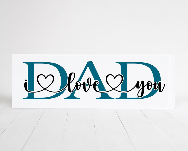 Puede incluir: Un letrero de madera blanco con la palabra "DAD" en letras azul turquesa. Las letras est&aacute;n conectadas por una l&iacute;nea negra con dos corazones. El texto "I love you" est&aacute; escrito en cursiva negra.