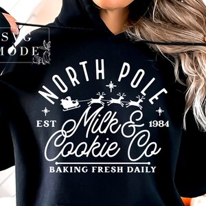 May include: Black hoodie with a white graphic design that says "North Pole Milk & Cookie Co. Est 1984 Baking Fresh Daily" with a silhouette of Santa Claus in a sleigh pulled by reindeer.