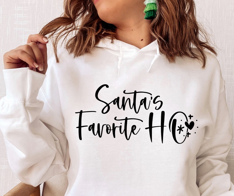 May include: White hooded sweatshirt with the text "Santa's Favourite Ho" in black cursive lettering. A small black heart with a snowflake and stars is next to the text.