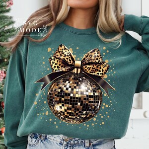 May include: A teal sweatshirt with a gold and black disco ball ornament with a leopard print bow. The ornament is surrounded by gold glitter.