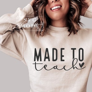 Op de afbeelding: Een beige sweatshirt met de tekst "MADE TO teach" in zwarte letters en een klein zwart hartje.