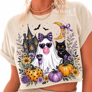 May include: A beige t-shirt featuring a graphic of a ghost wearing a purple bow and heart-shaped sunglasses, holding a coffee cup. The design includes a black cat, a haunted house, pumpkins, a crescent moon, and flowers in purple and orange.