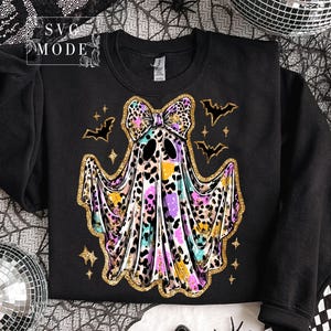 May include: Black sweatshirt featuring a colorful ghost design with a leopard print bow, outlined in gold glitter. The ghost has a watercolor effect with purple, yellow, and blue hues. Bats and stars complete the Halloween theme.