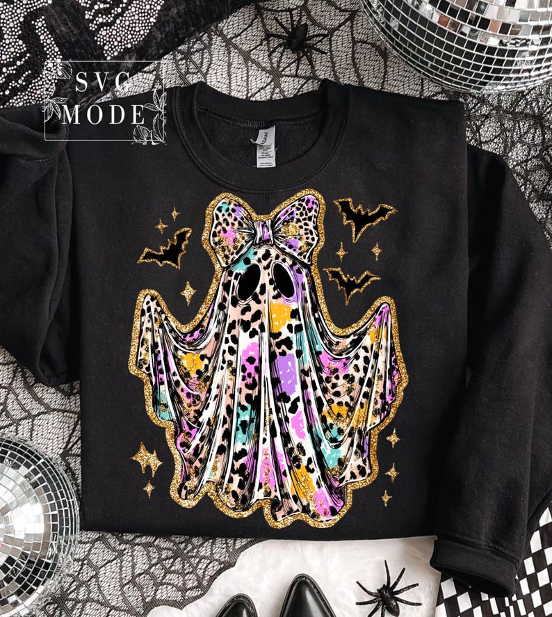 May include: Black sweatshirt featuring a colorful ghost design with a leopard print bow, outlined in gold glitter. The ghost has a watercolor effect with purple, yellow, and blue hues. Bats and stars complete the Halloween theme.