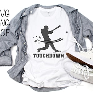 Puede incluir: Camiseta blanca con una silueta de un jugador de béisbol balanceando un bate con estrellas y el texto "TOUCHDOWN".