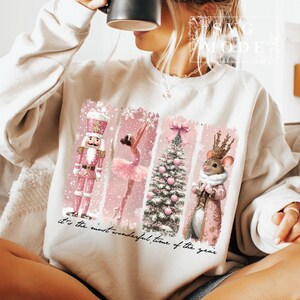 K&ouml;nnte beinhalten: Ein wei&szlig;er Pullover mit einem rosafarbenen Weihnachtsmotiv, das einen Nussknacker, eine Ballerina, einen Weihnachtsbaum und eine Maus zeigt. Der Text "it's the most wonderful time of the year" ist auf dem Pullover aufgedruckt.