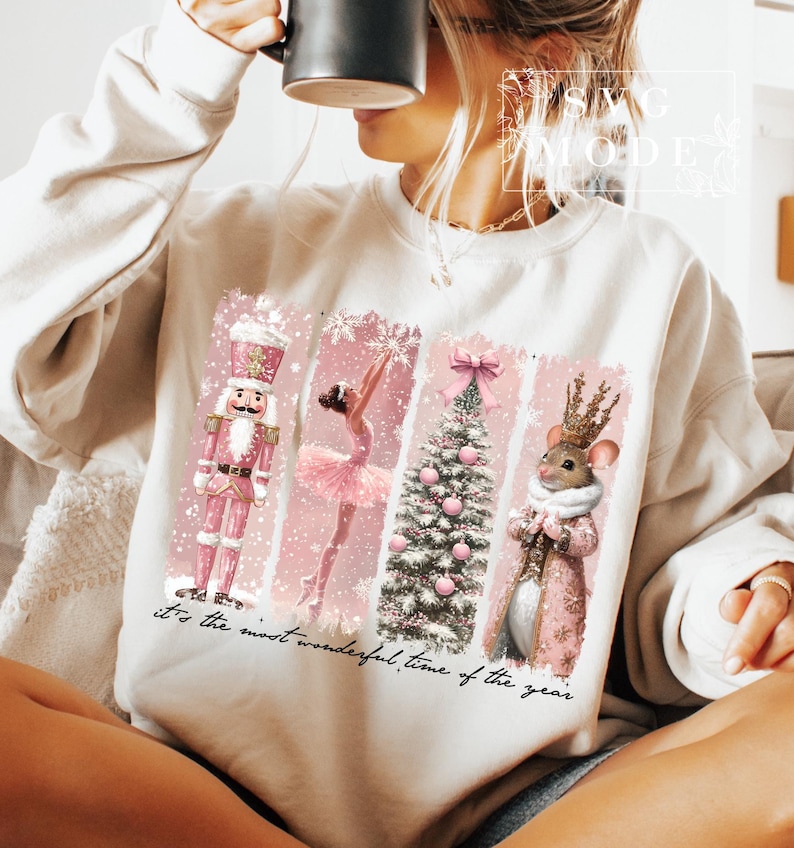 K&ouml;nnte beinhalten: Ein wei&szlig;er Pullover mit einem rosafarbenen Weihnachtsmotiv, das einen Nussknacker, eine Ballerina, einen Weihnachtsbaum und eine Maus zeigt. Der Text "it's the most wonderful time of the year" ist auf dem Pullover aufgedruckt.