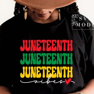 Può includere: Una maglietta nera con le parole "Juneteenth" ripetute tre volte in lettere rosse, verdi e gialle. Le parole "vibes" sono scritte in bianco con un cuore rosso.