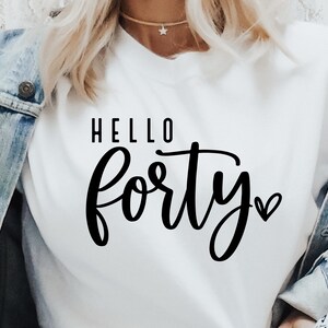 K&ouml;nnte beinhalten: Ein wei&szlig;es T-Shirt mit den Worten "HELLO Forty" in schwarzer Schrift. Ein kleines Herz befindet sich neben dem Wort "Forty". Das Shirt wird mit einer Jeansjacke getragen.