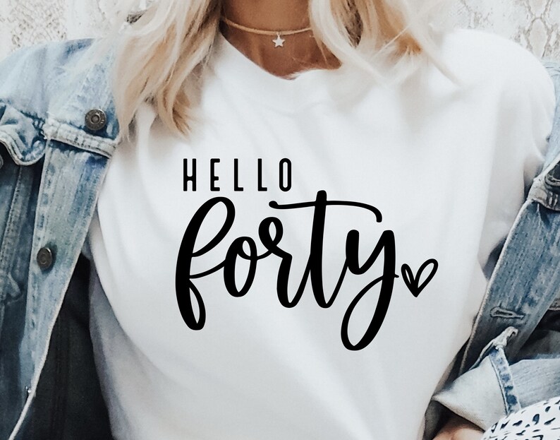 K&ouml;nnte beinhalten: Ein wei&szlig;es T-Shirt mit den Worten "HELLO Forty" in schwarzer Schrift. Ein kleines Herz befindet sich neben dem Wort "Forty". Das Shirt wird mit einer Jeansjacke getragen.