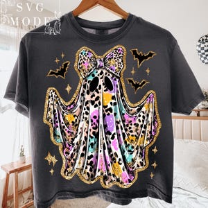 May include: Dark gray t-shirt featuring a colorful ghost design with a leopard print bow, gold glitter accents, and bat and star graphics. The shirt has a crew neck and short sleeves, perfect for Halloween.