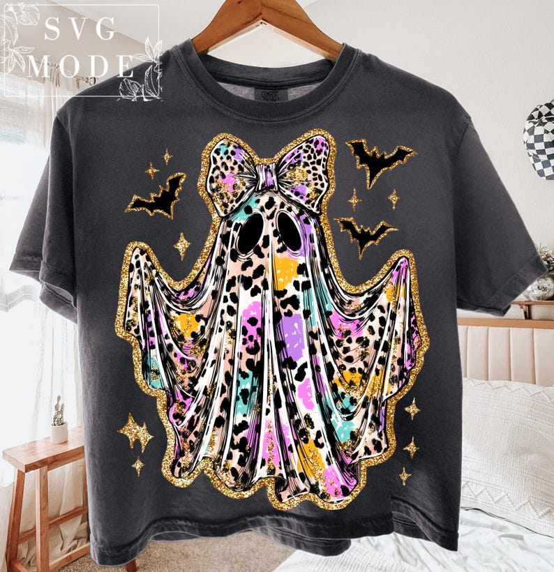 May include: Dark gray t-shirt featuring a colorful ghost design with a leopard print bow, gold glitter accents, and bat and star graphics. The shirt has a crew neck and short sleeves, perfect for Halloween.