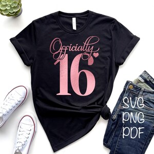 Puede incluir: Una camiseta negra con el texto "Officially 16" en letras de purpurina rosa. El n&uacute;mero 16 es grande y el texto "Officially" est&aacute; en una fuente cursiva. La camiseta est&aacute; colocada sobre una superficie blanca con un par de zapatillas blancas y unos vaqueros azules.
