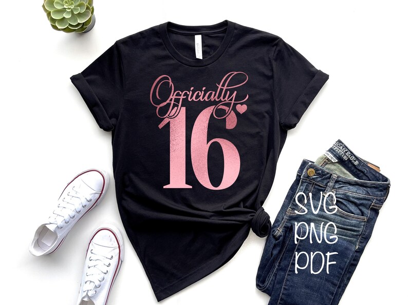 Puede incluir: Una camiseta negra con el texto "Officially 16" en letras de purpurina rosa. El n&uacute;mero 16 es grande y el texto "Officially" est&aacute; en una fuente cursiva. La camiseta est&aacute; colocada sobre una superficie blanca con un par de zapatillas blancas y unos vaqueros azules.