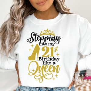 Pode incluir: Camisola branca com o texto "Stepping into my 21st Birthday like a Queen" em preto e dourado. O design inclui um salto alto, uma coroa e enfeites decorativos. Uma pe&ccedil;a de vestu&aacute;rio de estilo casual.