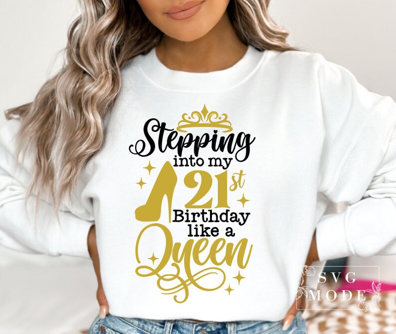 Pode incluir: Camisola branca com o texto "Stepping into my 21st Birthday like a Queen" em preto e dourado. O design inclui um salto alto, uma coroa e enfeites decorativos. Uma pe&ccedil;a de vestu&aacute;rio de estilo casual.
