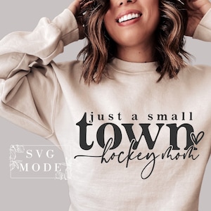 Op de afbeelding: Een beige sweatshirt met de tekst "just a small town hockey mom" in zwarte letters met een klein hartje.