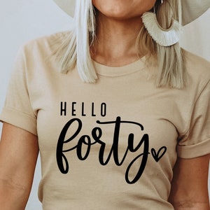 K&ouml;nnte beinhalten: Ein beiges T-Shirt mit hochgekrempelten &Auml;rmeln. Der schwarze Text lautet "HELLO forty" mit einem Herzsymbol. Das Shirt hat eine neutrale Farbe und einen l&auml;ssigen Stil.