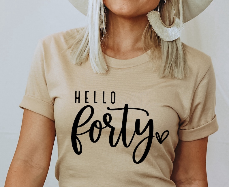 K&ouml;nnte beinhalten: Ein beiges T-Shirt mit hochgekrempelten &Auml;rmeln. Der schwarze Text lautet "HELLO forty" mit einem Herzsymbol. Das Shirt hat eine neutrale Farbe und einen l&auml;ssigen Stil.