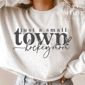 Op de afbeelding: Een grijs sweatshirt met de tekst "Just a small town hockey mom" in zwarte letters met een klein hartje.