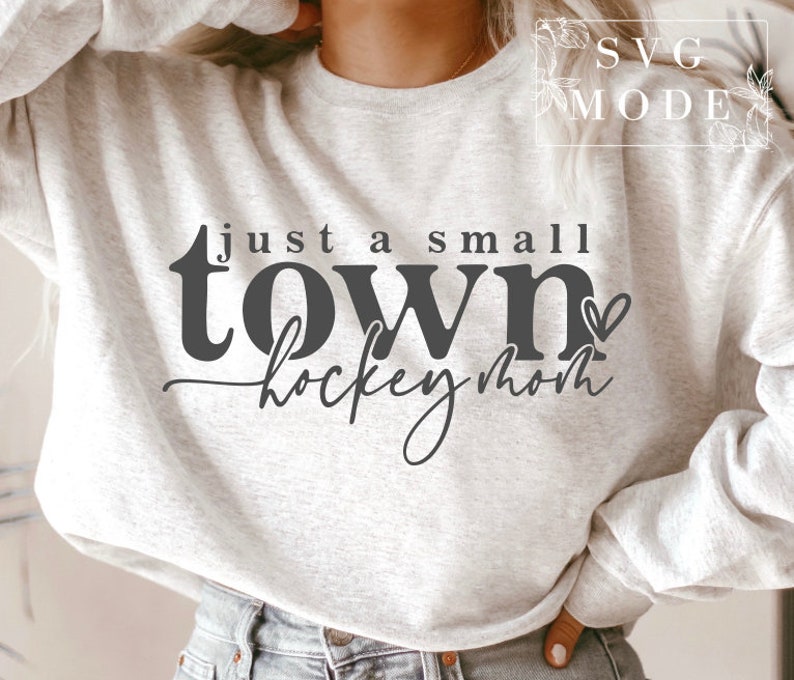 Op de afbeelding: Een grijs sweatshirt met de tekst "Just a small town hockey mom" in zwarte letters met een klein hartje.