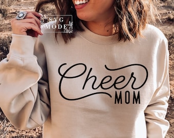 May include: A beige sweatshirt with the words "Cheer Mom" in black script. The sweatshirt has long sleeves and a crew neck. The text "SVG MODE" is in a white box in the upper left corner.