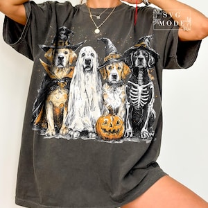 May include: Dark gray t-shirt featuring a Halloween-themed graphic. The design showcases four dogs dressed in costumes: a vampire, a ghost, a witch, and a skeleton, with a carved pumpkin in front. The text "SVG MODE" is in the upper right corner.