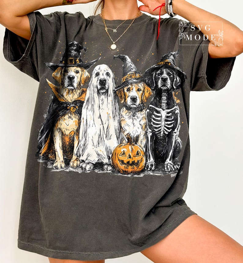 May include: Dark gray t-shirt featuring a Halloween-themed graphic. The design showcases four dogs dressed in costumes: a vampire, a ghost, a witch, and a skeleton, with a carved pumpkin in front. The text "SVG MODE" is in the upper right corner.