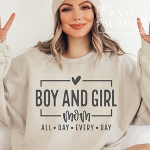 Könnte beinhalten: Ein beiger Pullover mit dem Text "Boy and Girl Mom" in schwarzer Schrift. Der Text ist von einem schwarzen Rechteck und einem kleinen Herzen darüber eingerahmt. Unter dem Text steht "All Day Every Day" in kleinerer schwarzer Schrift.