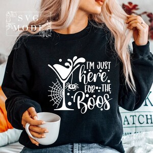 May include: Black sweatshirt with a white graphic design of a martini glass with a spider hanging from it and the text "I'm just here for the boos".