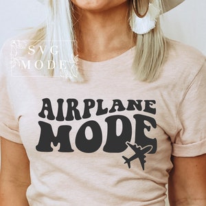 Puede incluir: Una camiseta rosa claro con el texto "AIRPLANE MODE" en letras negras de estilo retro. Un peque&ntilde;o gr&aacute;fico de avi&oacute;n est&aacute; debajo del texto.