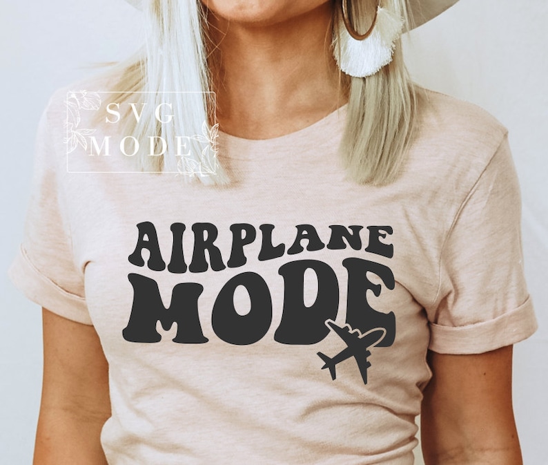 Puede incluir: Una camiseta rosa claro con el texto "AIRPLANE MODE" en letras negras de estilo retro. Un peque&ntilde;o gr&aacute;fico de avi&oacute;n est&aacute; debajo del texto.